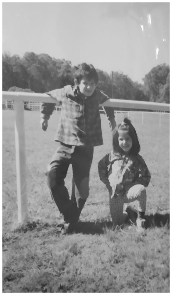 A black and white childhood photograph. It shows myself as a 8-year-old boy, wearing a plaid shirt and leaning casually against a white fence in a sunny, grassy field. My little 6-year-old sister is crouching happily next to me.
